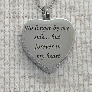 May include: A silver heart-shaped pendant necklace. The pendant has the engraved text "No longer by my side... but forever in my heart". The necklace is on a silver chain, and the background is a textured, off-white fabric.