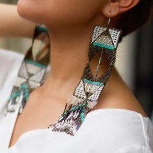 Peut inclure: Une paire de longues boucles d'oreilles en perles avec un motif géométrique dans des tons de brun, blanc et turquoise. Les boucles d'oreilles présentent une frange de perles en bas.