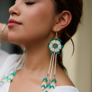 May include: A pair of long, beaded earrings with a white and teal flower design. The earrings are made of small, round beads and have a dangling, tassel-like design.