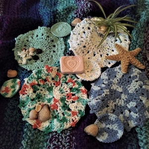 May include: A collection of crocheted dishcloths in various colours and patterns, including green, blue, white, and pink. There is a heart-shaped soap bar and seashells on a purple and blue patterned background.