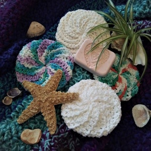 May include: A collection of six crocheted scrubbies in various colors, including white, pink, green, blue, and purple. The scrubbies are arranged on a purple and blue patterned fabric with a starfish and seashells.