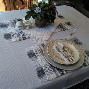 May include: A white tablecloth with two crocheted placemats, one with gray accents. A silver charger plate with a white plate and silverware sits on the gray placemat. A small potted plant with purple flowers sits in the center of the table.