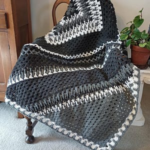May include: A handmade crochet blanket in shades of gray and white, draped over a wooden chair. The blanket features a textured pattern with white accents and a decorative border. This cozy throw is perfect for home decor.