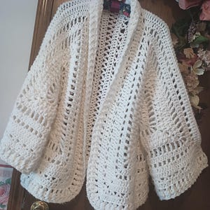 May include: A cream-colored, open-front crochet cardigan with a shawl collar. The handmade sweater features a textured, open weave design with three-quarter length sleeves. The cardigan is hanging on a wooden door.