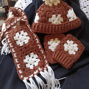 May include: A brown crocheted hat with a white pom pom, matching scarf and fingerless gloves. The hat, scarf, and gloves are all made with a white and brown granny square pattern.