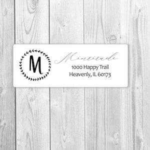 Puede incluir: Una etiqueta de dirección de devolución blanca con una corona negra y la letra "M" en el centro. El texto "Mintz" está escrito en cursiva seguido de la dirección "1000 Happy Trail, Heavenly, IL 60173".