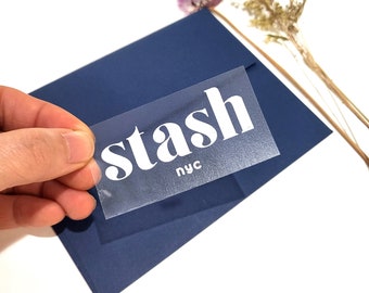 Könnte beinhalten: Ein transparenter Aufkleber aus Plastik mit dem Wort "stash" in weißen Fettdruckbuchstaben und "nyc" in kleineren weißen Buchstaben darunter. Der Aufkleber befindet sich auf einem blauen Hintergrund.