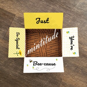 May include: A yellow and white gift box with a honeycomb pattern and the text "Just You're Bee-cause" on the front. The box is open and the inside is lined with brown paper. The sides of the box have the text "So Special" and "mintitude" on them.