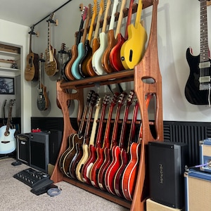 Può includere: Un supporto per chitarra in legno con 18 chitarre elettriche, alcune con finiture rosse, gialle, blu e bianche. Il supporto è in una stanza con una parete a motivi bianco e nero e un altoparlante nero.