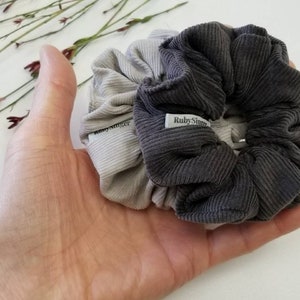 May include: Two fabric scrunchies in gray and beige colors. The scrunchies are made of corduroy fabric and have a small label that says "Ruby Singer".