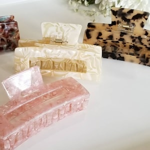 以下が含まれることがあります： 異なる色と模様のプラスチック製のヘアクリップが4つあります。クリップはすべて同じサイズと形をしていて、長方形のベースと湾曲したトップを持っています。クリップは半透明の素材で作られており、茶色、白、ピンク、黒など、さまざまな色と模様があります。