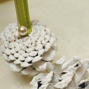 May include: Two white painted pine cones with glitter, each with a white pearl bead attached to a green ribbon.