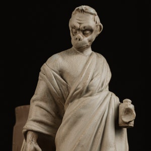 Può includere: Una statua in ceramica bianca di una persona con un viso simile a quello di una scimmia, che indossa una toga e tiene una lanterna. La statua è incisa con il testo "DIOGENE NE S".