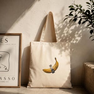 Peut inclure: Un sac fourre-tout en toile couleur crème arborant un motif central représentant une banane jaune fixée par un morceau de ruban adhésif gris argenté. Le sac possède deux longues anses et une forme rectangulaire en tissu tissé.