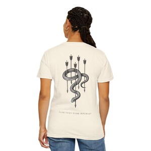 May include: A cream-coloured short-sleeve t-shirt featuring a black graphic on the back. The design depicts a coiled snake intertwined with five vertical arrows. Below the illustration, the text reads: FLUX TUUS VIAM INVENIAT.