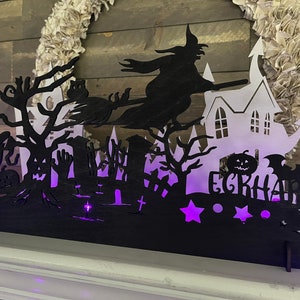 May include: Black wooden Halloween silhouette with a witch flying on a broomstick, a haunted house, a pumpkin, and the text "EGRHARD" in a spooky font. The silhouette is lit with purple lights.