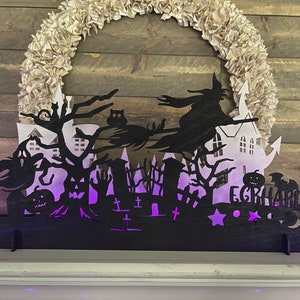 May include: Black silhouette of a witch flying on a broomstick over a spooky Halloween scene with a haunted house, trees, and tombstones. The scene is illuminated with purple lights and the name "E.GRAHARD" is visible at the bottom.