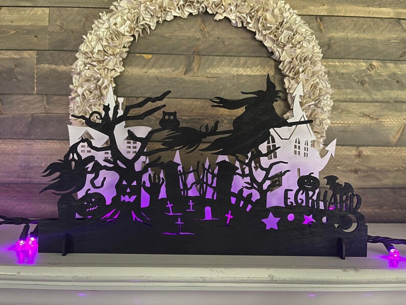 May include: Black silhouette of a witch flying on a broomstick over a spooky Halloween scene with a haunted house, trees, and tombstones. The scene is illuminated with purple lights and the name "E.GRAHARD" is visible at the bottom.
