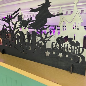 May include: Black and white Halloween-themed silhouette decoration with a witch flying on a broomstick, a haunted house, and a pumpkin. The decoration has the name "EGRHARD" written on it.