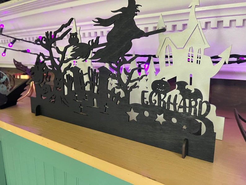 May include: Black and white Halloween-themed silhouette decoration with a witch flying on a broomstick, a haunted house, and a pumpkin. The decoration has the name "EGRHARD" written on it.