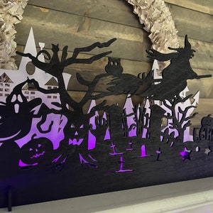 May include: Black silhouette Halloween decoration with a witch flying on a broomstick, a ghost, a haunted house, and a graveyard. The decoration is lit from behind with purple light.