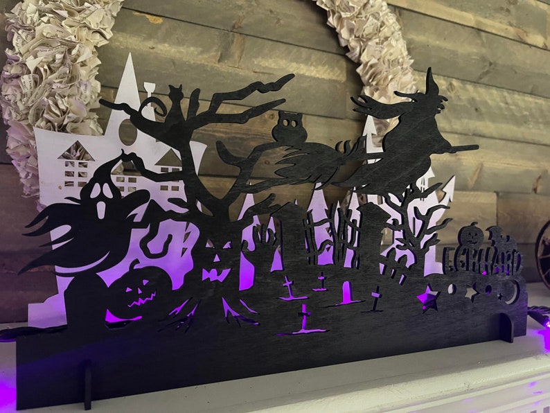 May include: Black silhouette Halloween decoration with a witch flying on a broomstick, a ghost, a haunted house, and a graveyard. The decoration is lit from behind with purple light.