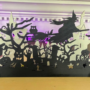 May include: Black wooden Halloween silhouette with a witch flying on a broomstick, a haunted house, trees, and pumpkins. The text "Eerie-hard" is at the bottom of the silhouette.