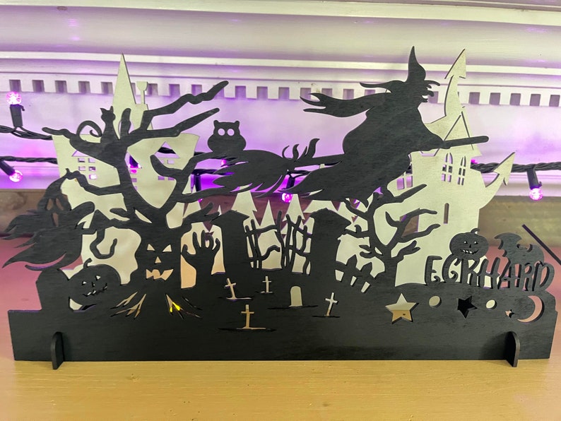 May include: Black wooden Halloween silhouette with a witch flying on a broomstick, a haunted house, trees, and pumpkins. The text "Eerie-hard" is at the bottom of the silhouette.
