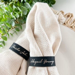 May include: Two beige linen napkins with black leather napkin rings. The rings are personalized with the names "Charlie John" and "Abigail Young" in gold lettering.