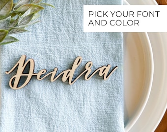 May include: A wooden place card with the name "Deidra" written in a cursive font. The place card is on a light blue napkin and a white plate. The text "PICK YOUR FONT AND COLOR" is visible in the top right corner of the image.