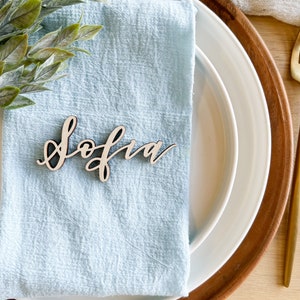 May include: A light blue napkin with a wooden place card that says "Sofia" on it. The place card is on a white plate with a brown wooden charger.