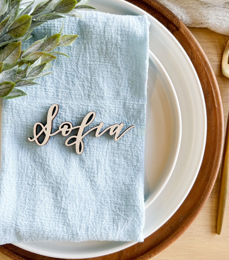 May include: A light blue napkin with a wooden place card that says "Sofia" on it. The place card is on a white plate with a brown wooden charger.