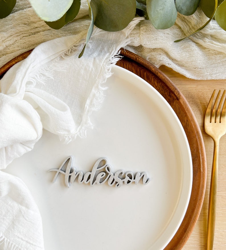 May include: A silver-colored wooden place card with the name "Anderson" written in cursive script. The place card is sitting on a white plate with a white napkin.
