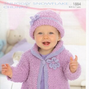 May include: A purple knitted baby cardigan with a flower detail and a matching purple hat. The cardigan has a button closure and a collar. The hat has a bow on top. The pattern is called "Snuggly Snowflake Chunky" and the number is 1884. Birth to 7 years.