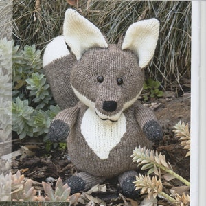 May include: A knitted fox toy with brown and white fur. The fox is sitting on a bed of brown leaves and green plants. The text "Fox" is printed below the toy. The text "Wool Blend DK" is printed below the text "Fox".