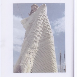 May include: A cream-coloured, hand-knitted blanket with a woven pattern and fringe. The blanket is draped over a person against a backdrop of a blue sky with clouds. The blanket's texture is visible.