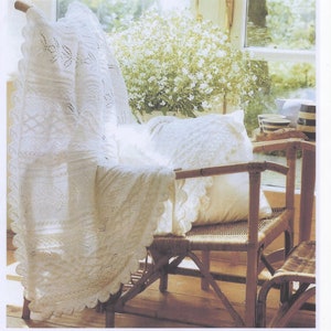 May include: A white crocheted baby blanket draped over a wicker chair with a white cushion. The chair is in a sunlit room with a potted plant in the background.