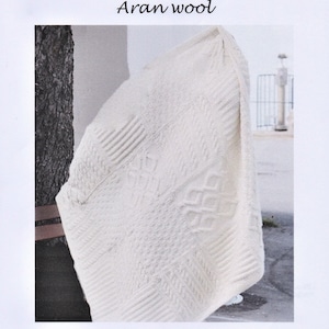 Puede incluir: Una manta Aran de color blanquecino hecha de lana Aran. La manta presenta una variedad de patrones de punto texturizados, incluyendo diseños de cable y diamante. El texto "Aran Blanket" y "Aran wool" es visible en la parte superior de la imagen.