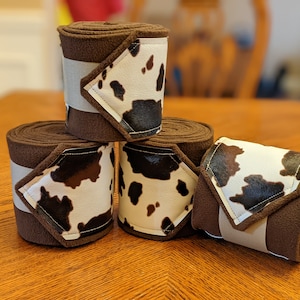 Può includere: Quattro fasce marroni per cavalli con dettagli a stampa di mucca. Ogni fascia è arrotolata e fissata con una cinghia grigio chiaro. La stampa della mucca è bianca con macchie nere e marroni. Le fasce sono impilate su una superficie di legno.