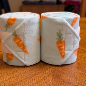 May include: Two rolled-up, light blue fleece wraps with orange carrot designs. Each wrap is secured with a white strap. The wraps are on a wooden surface. The carrot design includes green tops.