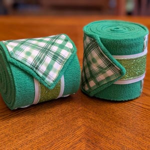 Puede incluir: Dos vendas verdes enrolladas para las patas de los caballos. Cada venda presenta un acento a cuadros verdes y blancos, una banda verde brillante y una cinta blanca. Las vendas se muestran sobre una superficie de madera.