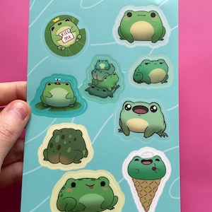 May include: A sheet of stickers featuring various cartoon frogs in different poses. The frogs are green with black eyes and some have a crown, a fly, or ice cream. The stickers are on a light blue background with white lines.