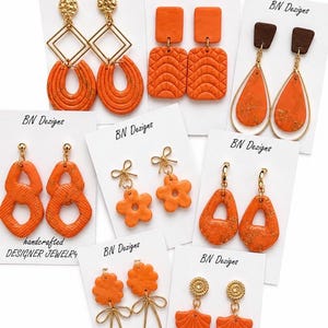 May include: A collection of eight pairs of orange earrings displayed on white cards labeled BN Dezigns. The earrings feature various shapes including hoops, rectangles, teardrops, flowers, and shells, accented with gold-tone metal and textured details.