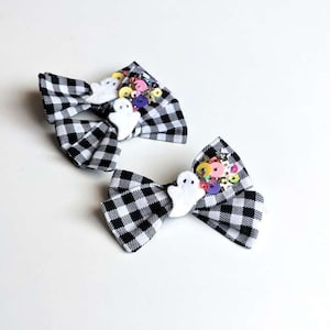 May include: Two black and white checkered hair bows with white ghost embellishments and colorful buttons. The bows are perfect for Halloween.