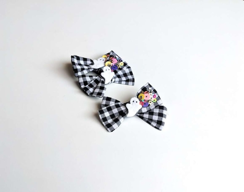 May include: Two black and white checkered hair bows with white ghost embellishments and colorful buttons. The bows are perfect for Halloween.