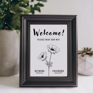 Puede incluir: Un cartel enmarcado en negro con fondo blanco y el texto "¡Bienvenido! Disfruta de nuestra wifi" con una ilustración en blanco y negro de dos flores. El cartel también incluye el nombre de la red "TheJohnsons" y la contraseña "password123".