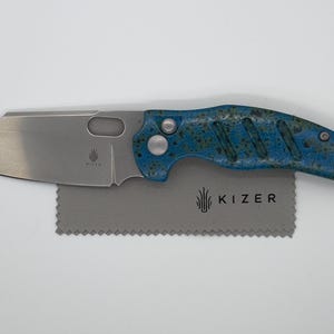 May include: A silver folding knife with a blue and green speckled handle. The blade is partially open, and the knife rests on a gray cloth with the brand name "KIZER" printed on it. The knife has a unique, modern design.