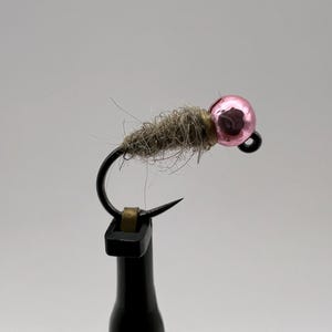 May include: A pink bead fly fishing lure with a brown and tan body. The lure is tied to a black hook and is mounted on a black fly tying vise.