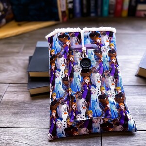 May include: A purple and blue fabric phone case with a white fuzzy lining. The case features a repeating pattern of characters from the animated film Frozen. The case has a pocket for a phone and a button closure.
