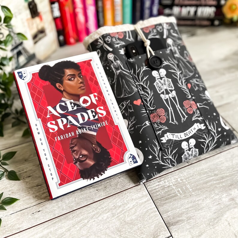 May include: A paperback book titled "Ace of Spades" by Faridah Abike-Inimide. The book cover features a red background with a black and white illustration of a person wearing a black and white shirt. The book is lying on a wooden surface with a black and white book cover with a floral design.